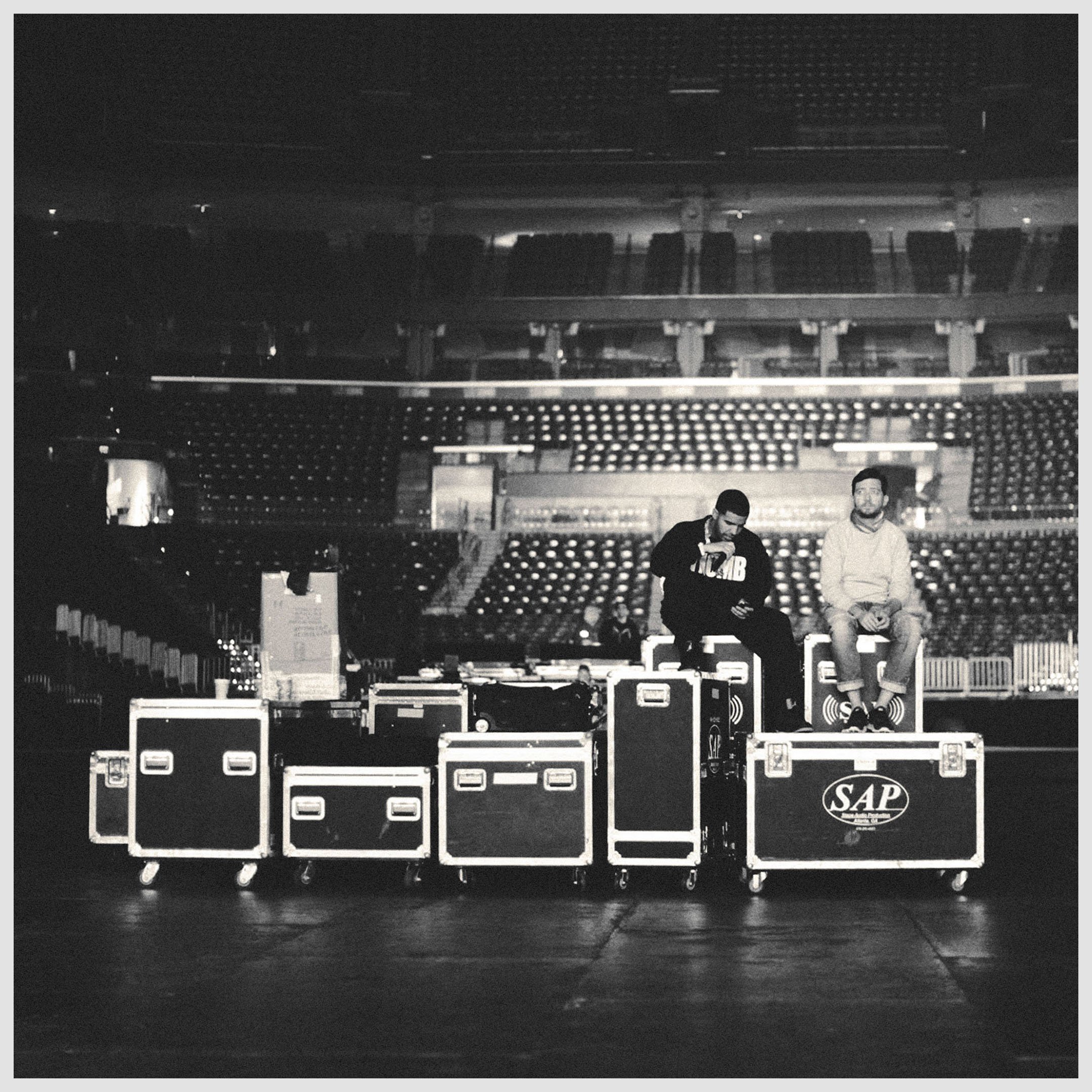 Black and white shot of two adult males sitting on road cases inside a stadium with rows of empty seating in the background.
