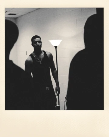 Black and white shot of adult male in a tank top with necklaces, facing forward while two other adults stand in the foreground.