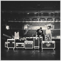 Black and white photo of Drake and a companion seated on stage flight cases in an empty arena bowl.