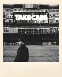 Monochrome Polaroid photograph of Drake standing in a stadium with a giant Take Care sign overhead.