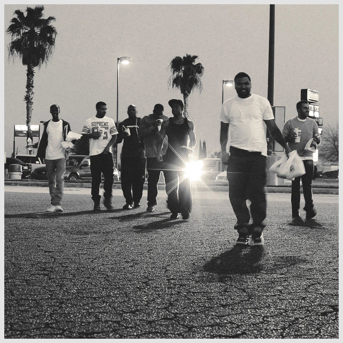 Group of men walking across a paved parking lot at night with palm trees in the background under streetlights.