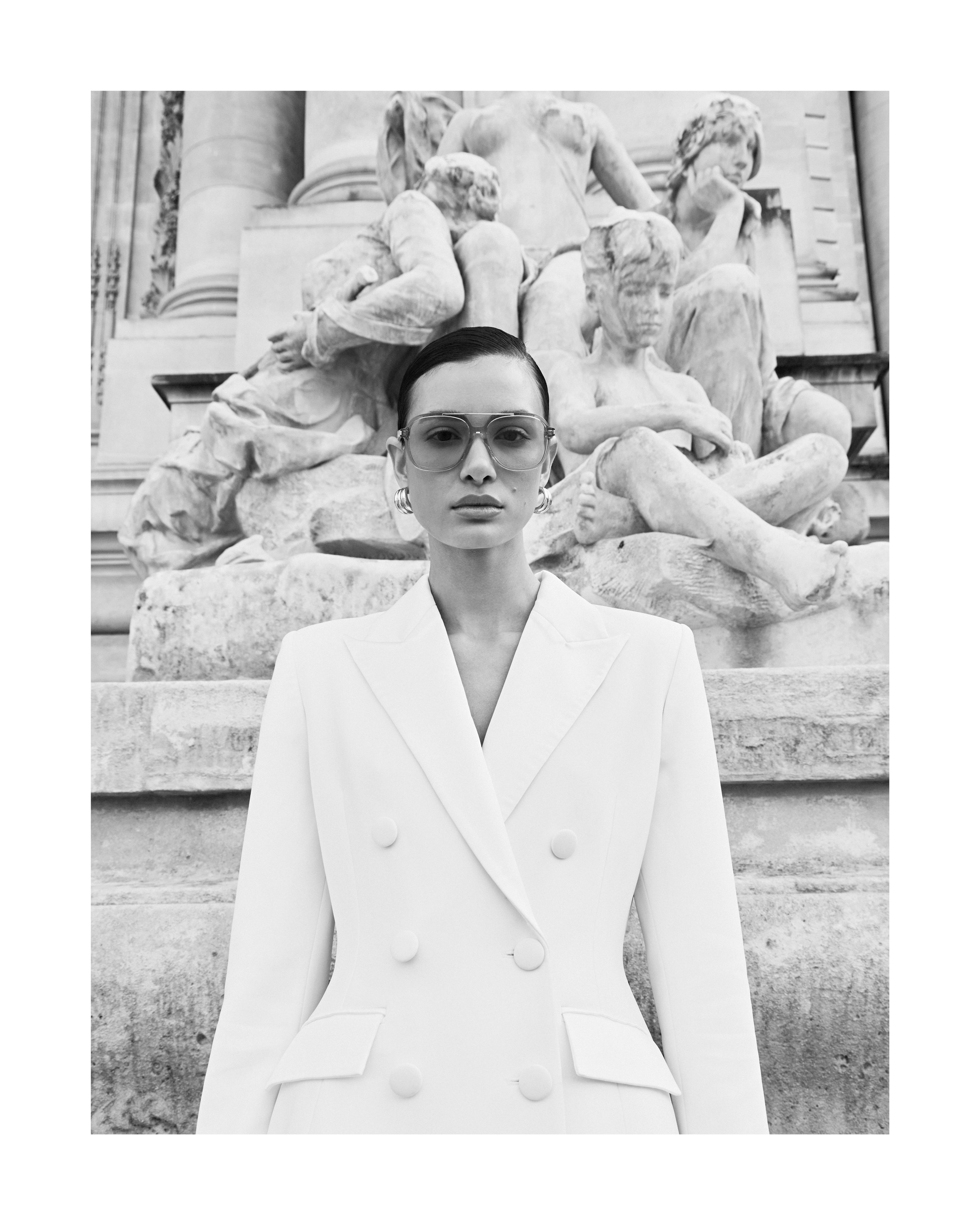 A woman poses outdoors in a white double-breasted blazer and sunglasses, standing in front of a weathered stone sculpture in a fashion editorial.