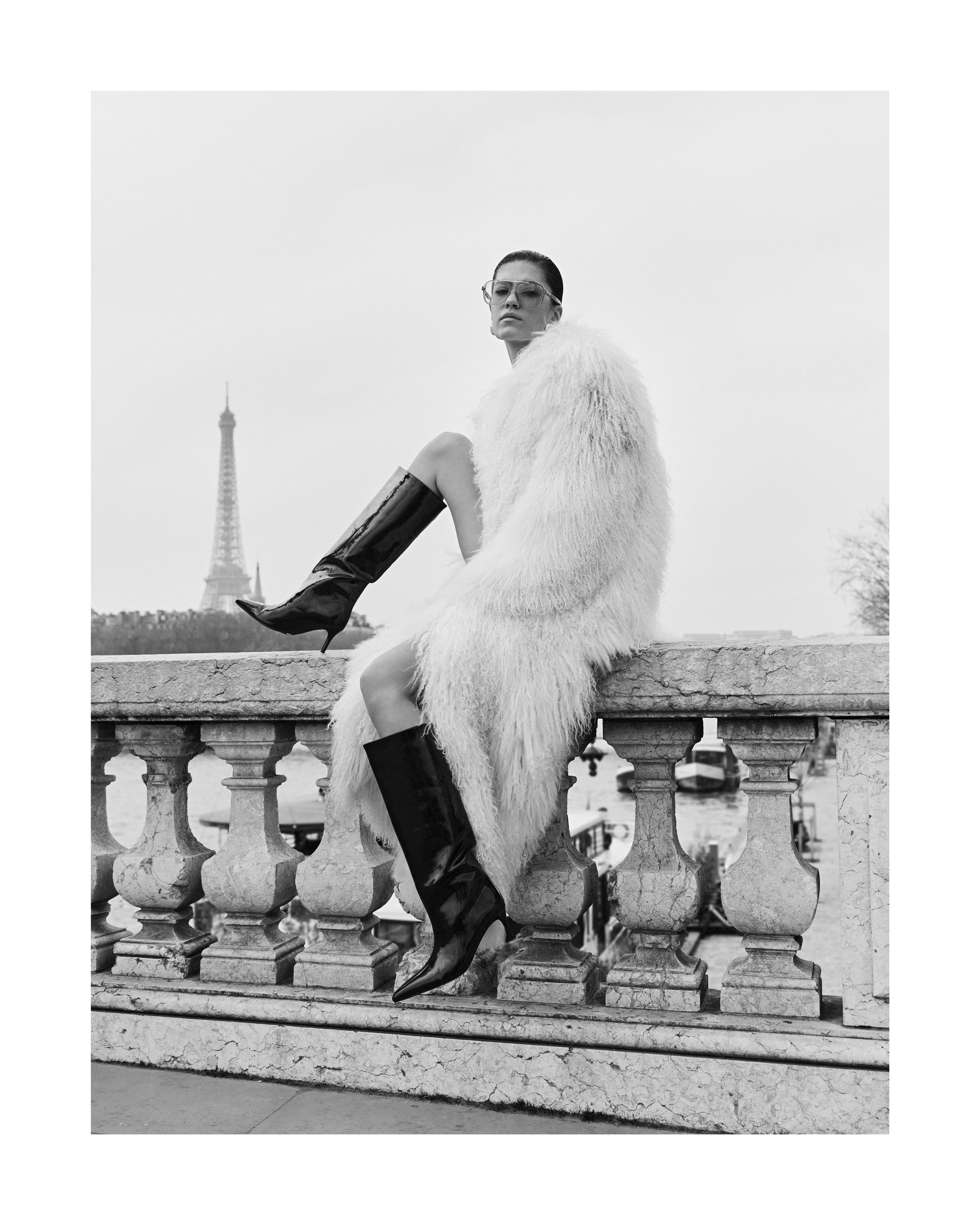Model wearing a white shaggy coat and tall black boots sitting on a stone balustrade with the Eiffel Tower in the background