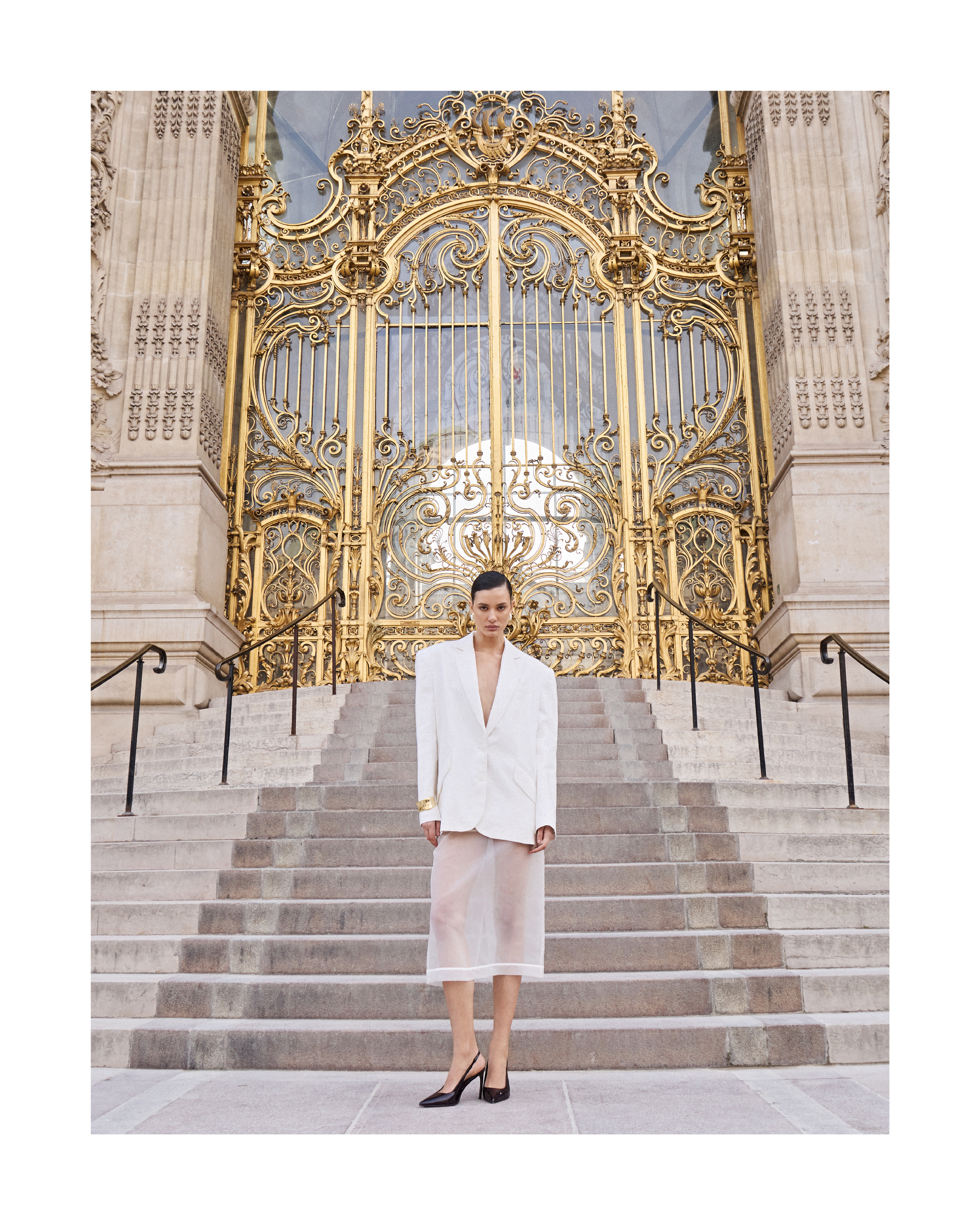 Elegant woman in white blazer and sheer skirt poses on stone steps before ornate golden gates, fashion photography.