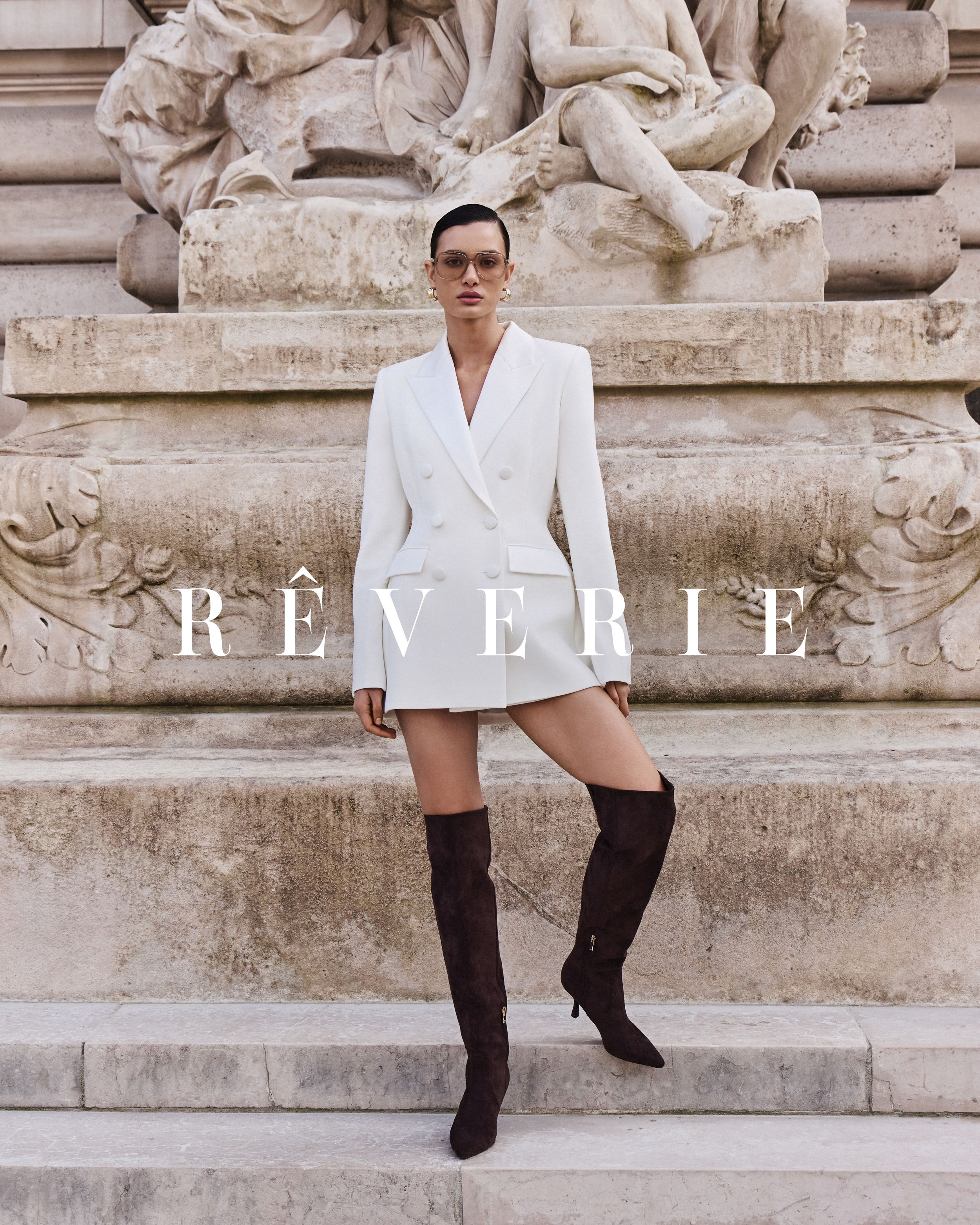 Fashionable woman in white blazer dress and brown thigh-high boots poses in front of a weathered stone structure with a sculpture.