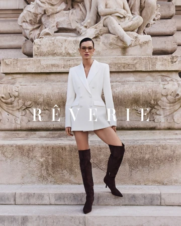 Fashionable woman in white blazer dress and brown thigh-high boots poses in front of a weathered stone structure with a sculpture.