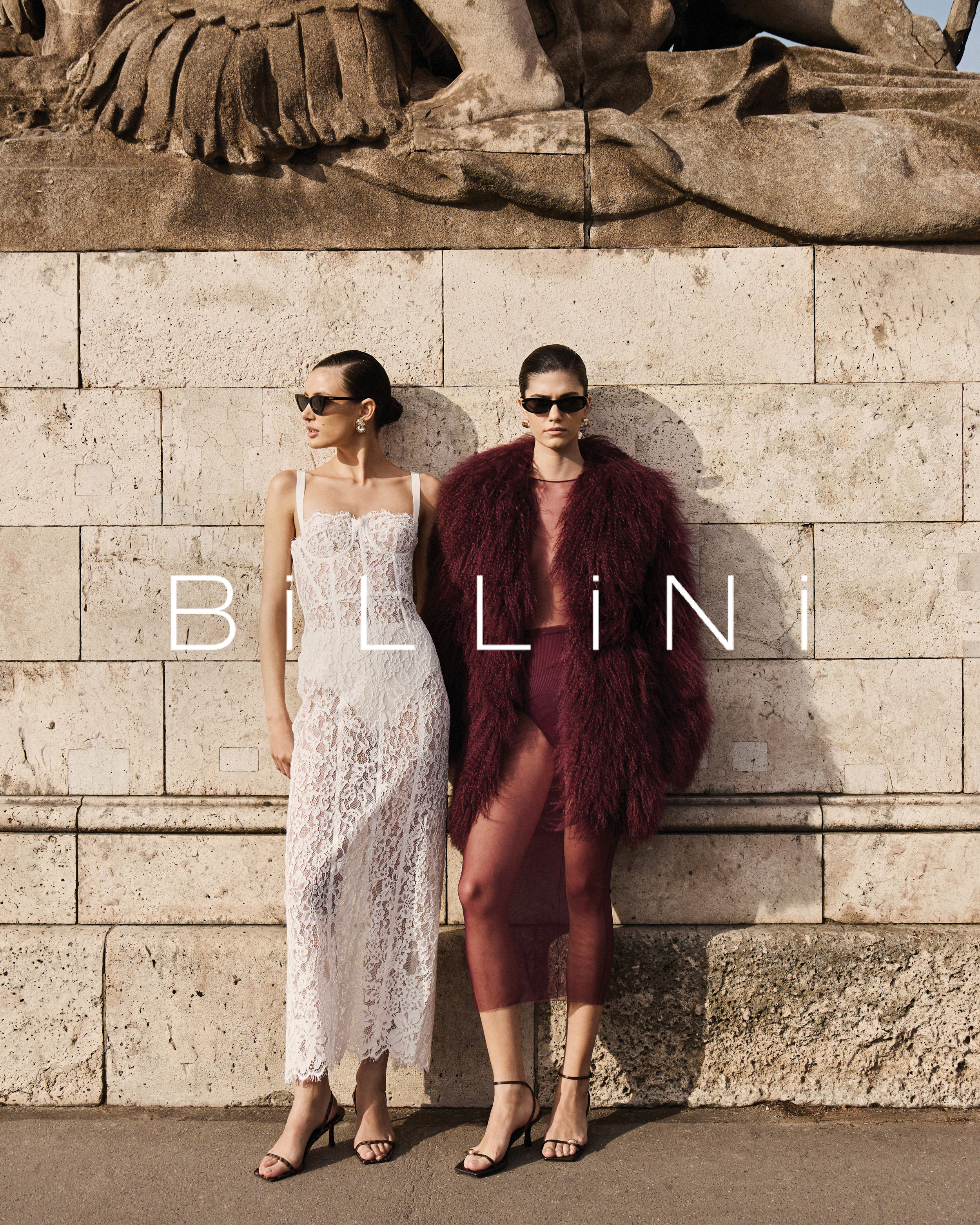 Two fashion models pose outdoors near a stone wall wearing dresses and heels. The models are wearing sunglasses and fashionable attire.