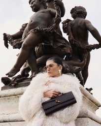 Woman in white fuzzy coat holding a dark woven clutch in front of stone statues
