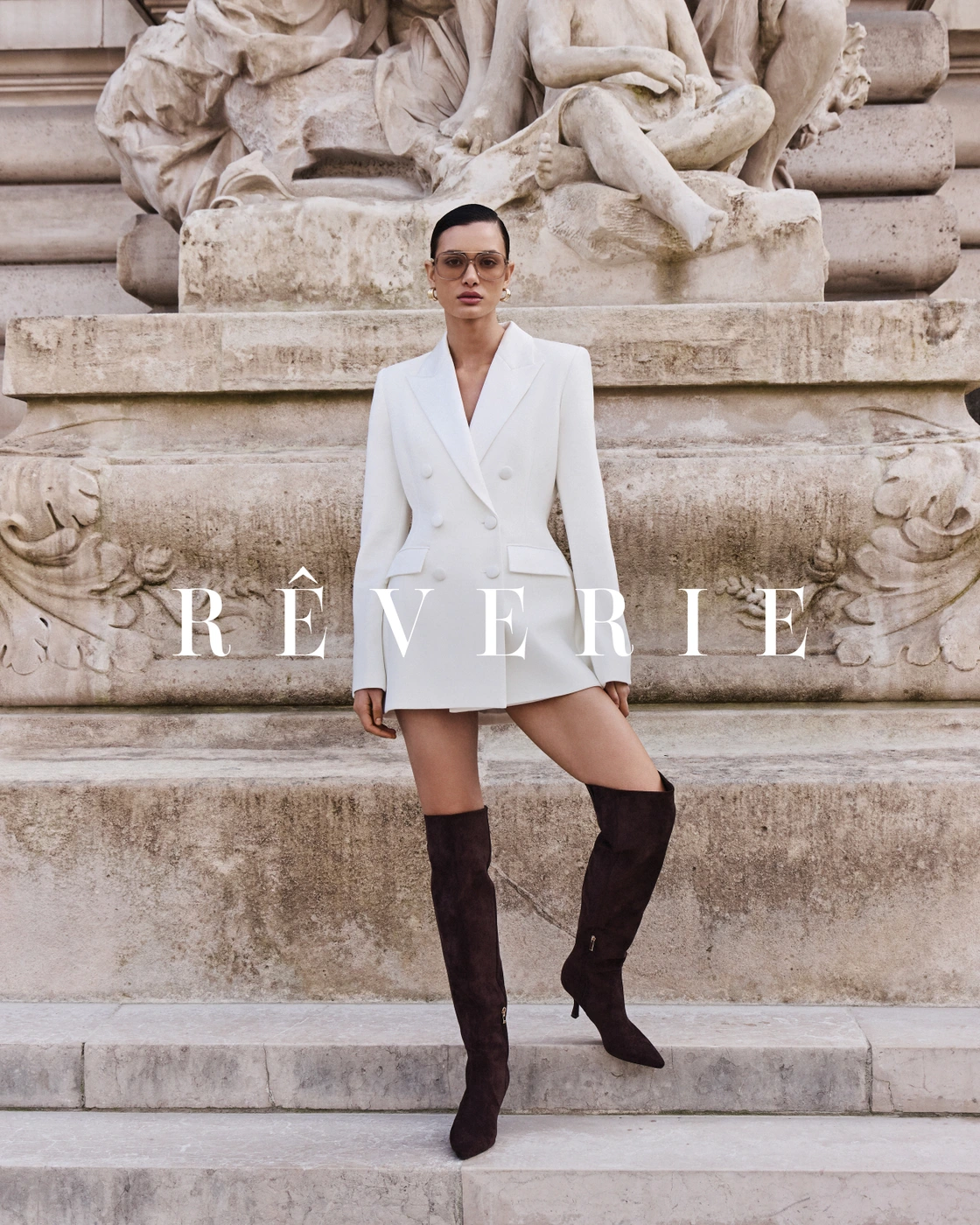 Model wearing a white double-breasted blazer and dark brown over-the-knee boots posing in front of a classical stone monument.