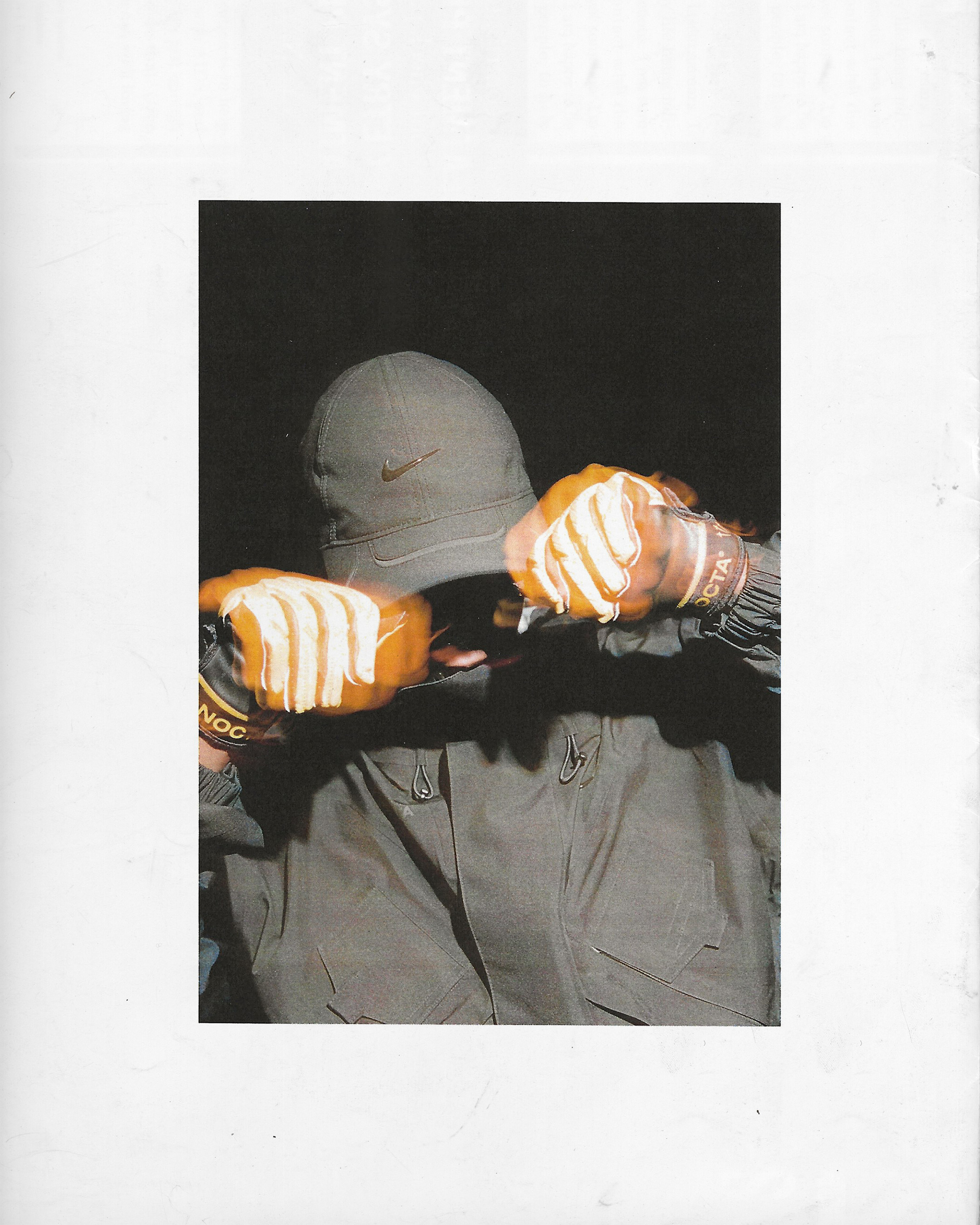 A person in an olive-green jacket and cap covers their face with gloved hands against a dark background. Part of the torso visible.
