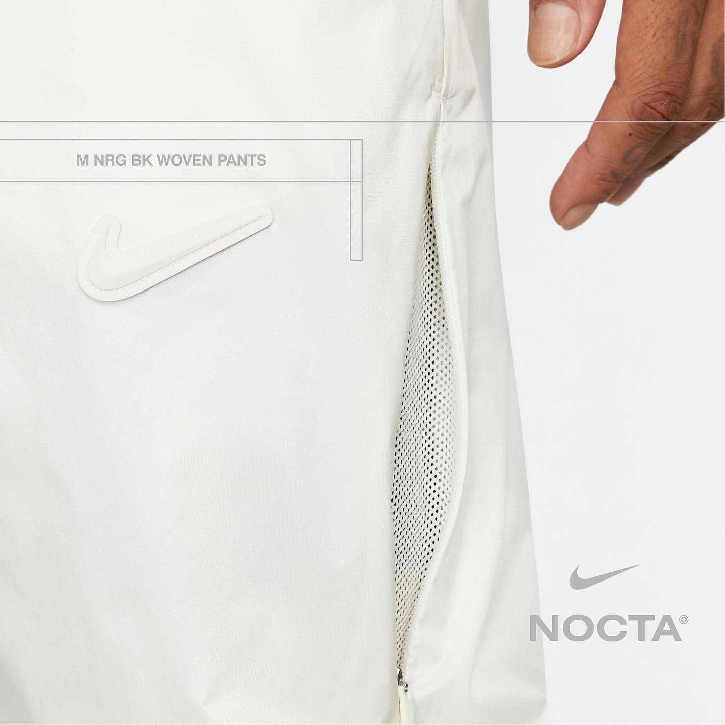 Close-up of cream-colored Nike pants featuring the Nike swoosh and "NOCTA" logo, a side zipper pocket with mesh, and an adult hand.