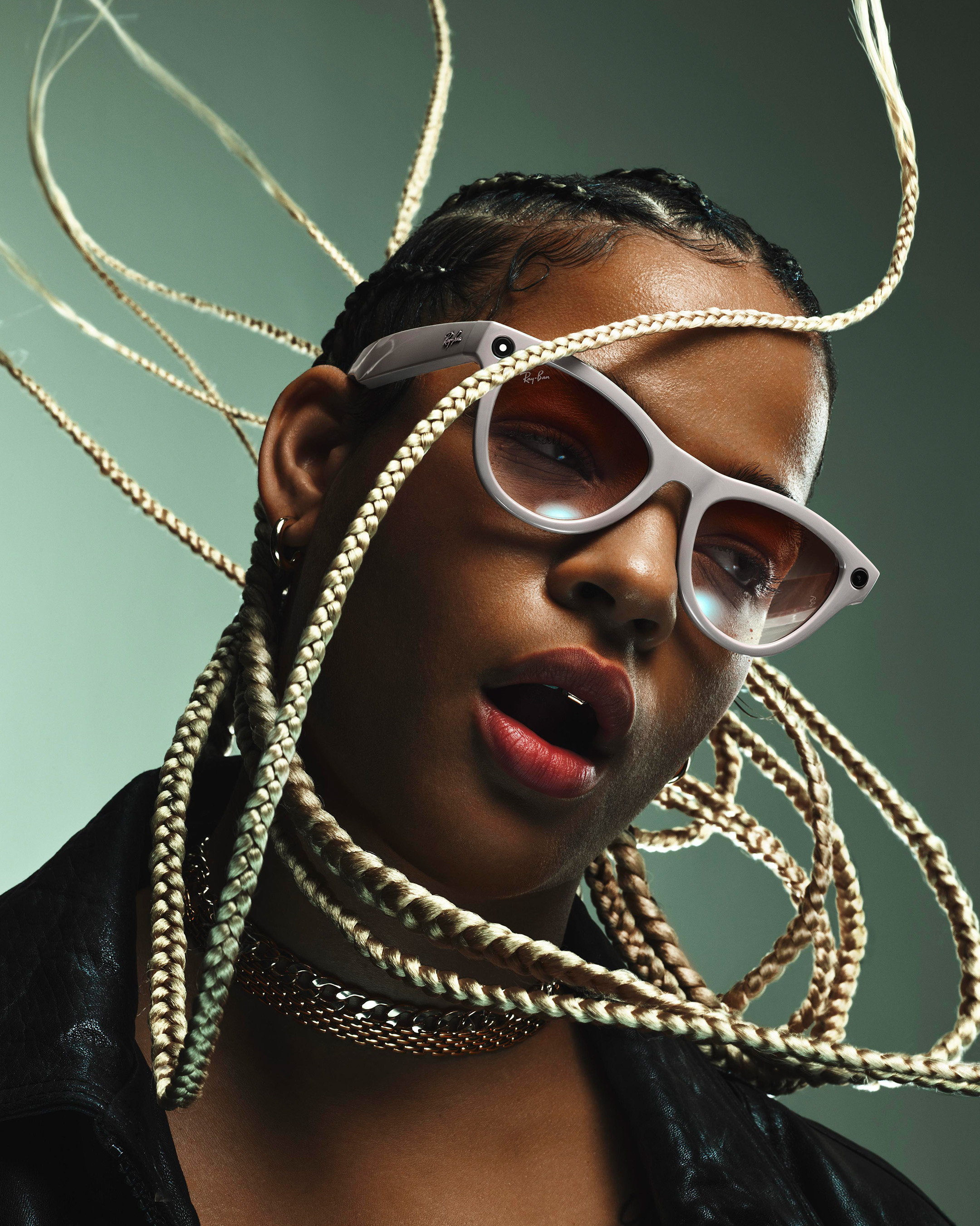 Close-up studio shot of a person with dark skin, blonde braids, gold chain, wearing white-rimmed smart glasses, against a green background.