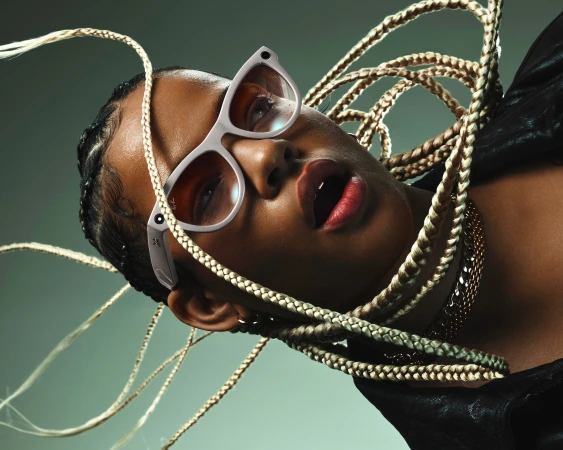 Close-up studio shot of a person with dark skin, blonde braids, gold chain, wearing white-rimmed smart glasses, against a green background.