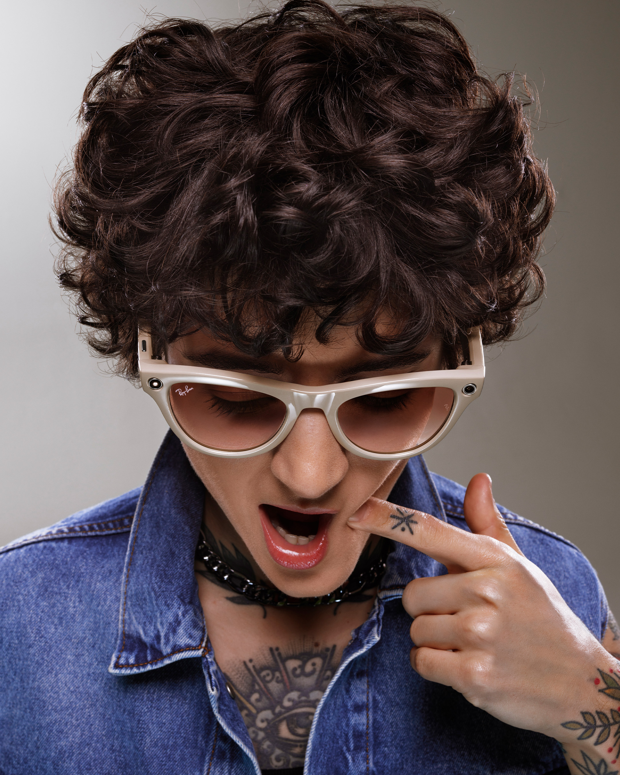 Person with curly hair, sunglasses, and tattoos, wearing a denim jacket, poses with mouth open and finger pointing to their cheek.