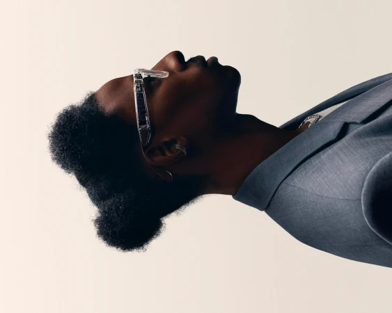 Portrait of a Black man in a gray suit and clear glasses looking up against a cream background in a brightly lit, high-angle shot.