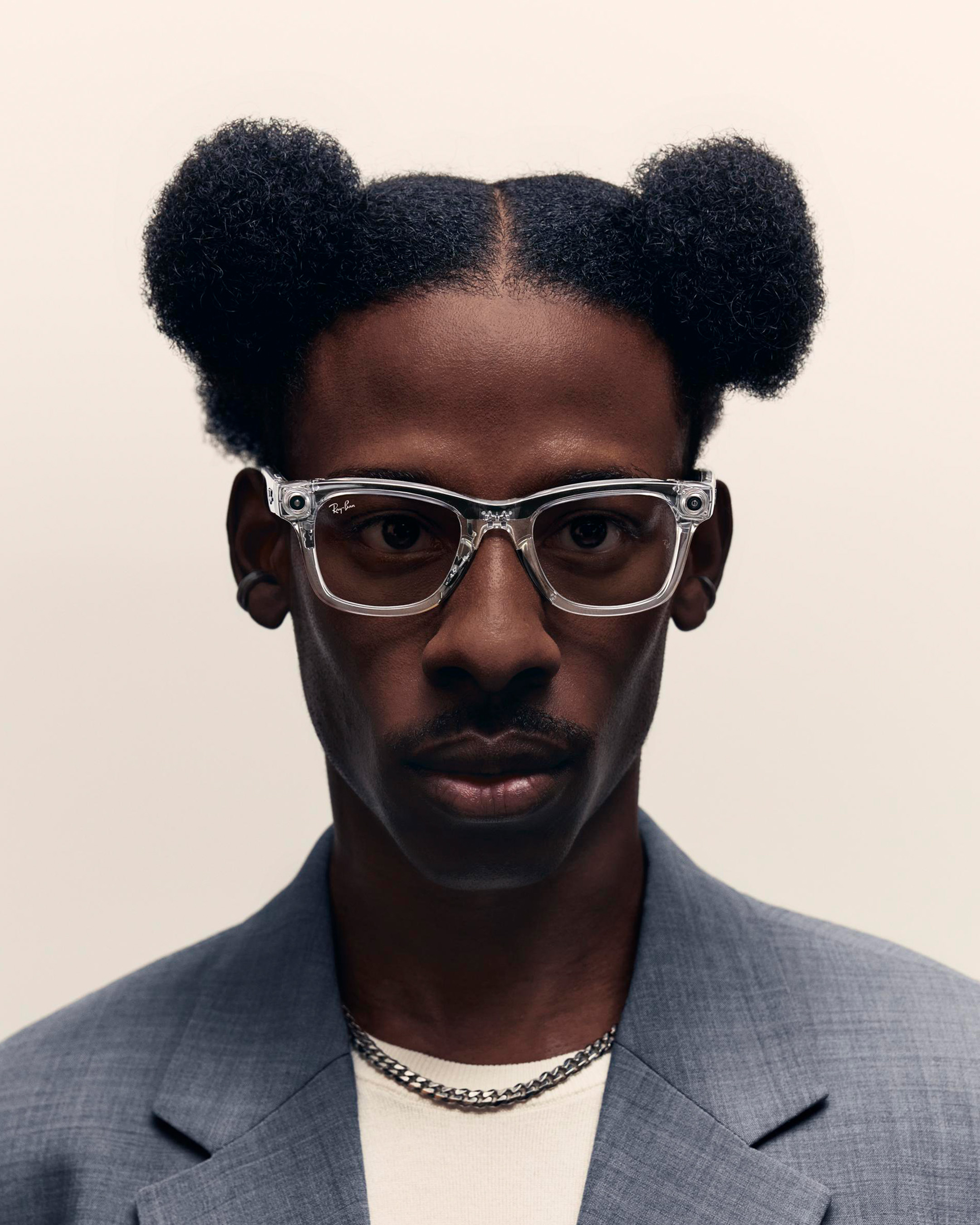 Close-up of a Black man wearing clear Ray-Ban glasses and a grey suit, posing against an off-white backdrop in a portrait shot.
