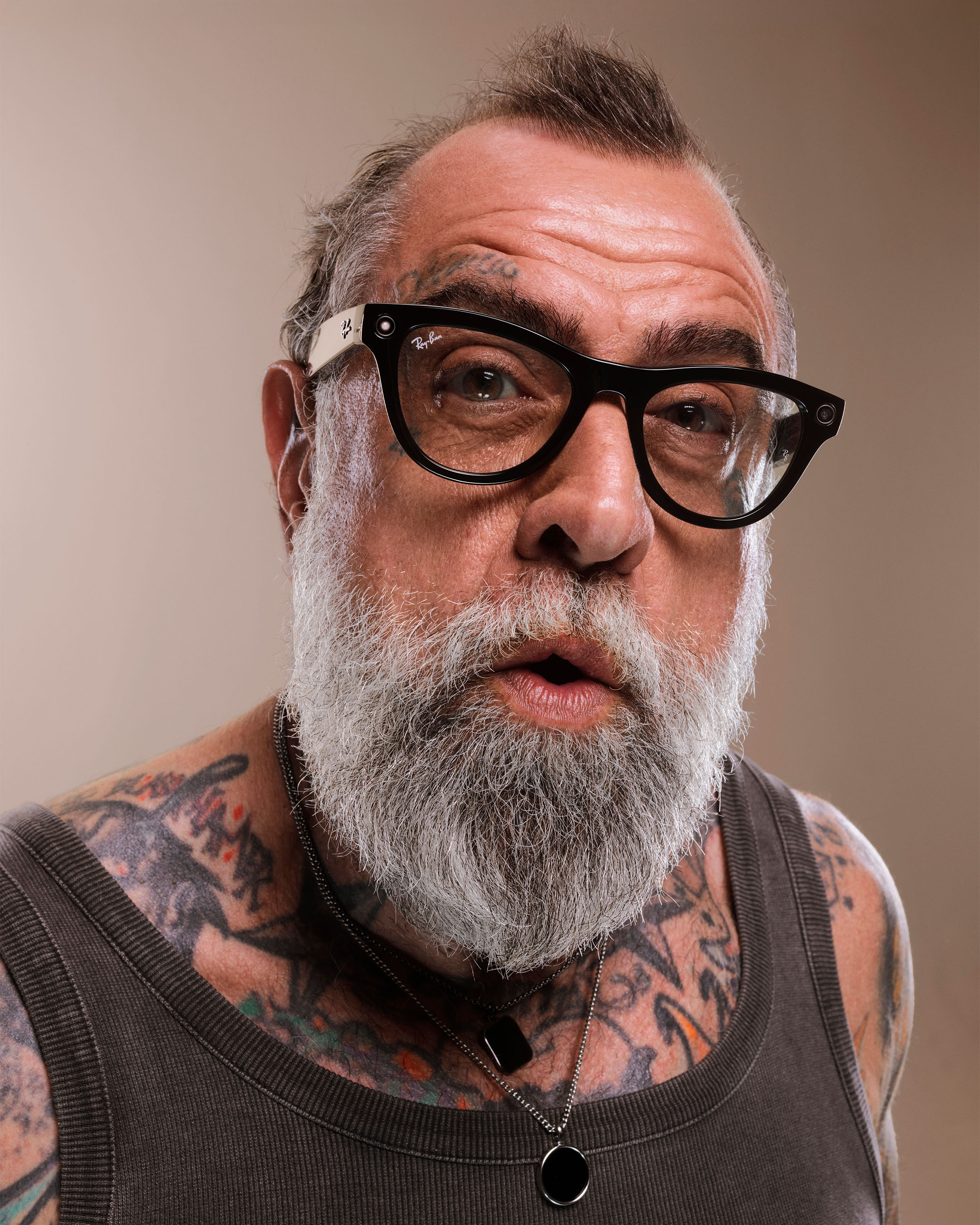 Portrait of a mature man with gray beard, tattoos, and Ray-Ban glasses looking directly at the viewer against a neutral background.
