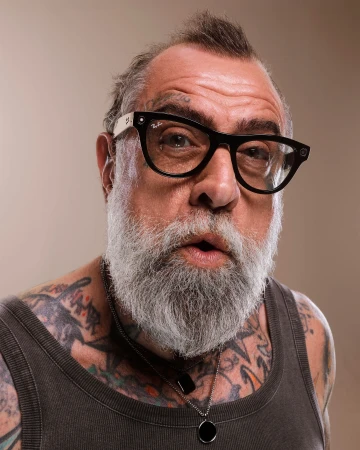 Portrait of a mature man with gray beard, tattoos, and Ray-Ban glasses looking directly at the viewer against a neutral background.
