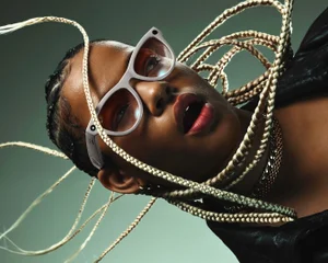 Close-up portrait of a woman wearing smart glasses with light frames and blonde braids framing her face against a green gradient background.
