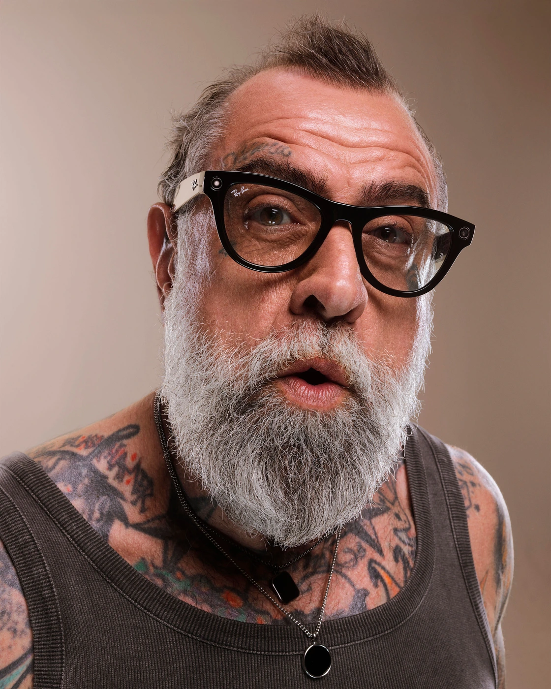 Tattooed man with a full grey beard wearing black smart glasses and a dark ribbed tank top against a warm neutral studio backdrop.