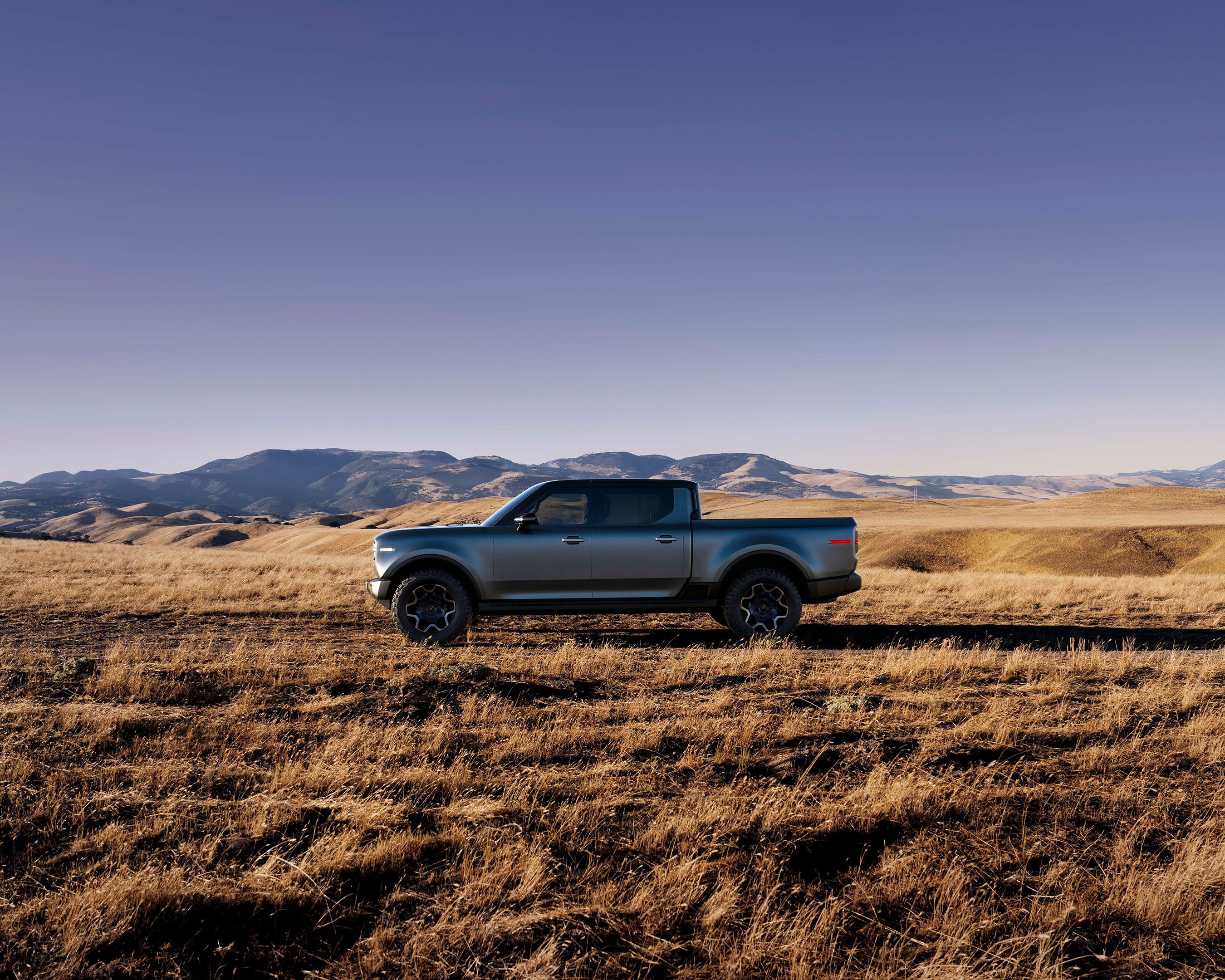 Silver pickup truck parked in a field with mountains in the background under a blue sky. Automotive vehicle, rural environment.