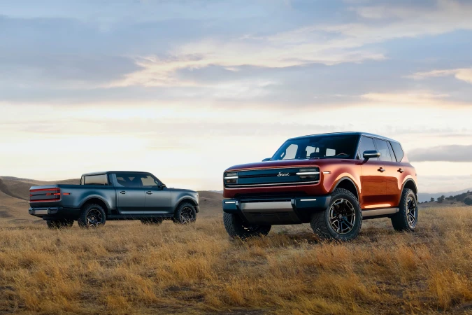 Two electric vehicles, a pickup truck and SUV, are parked on a grassy field under a sky with wispy clouds at sunset or sunrise.