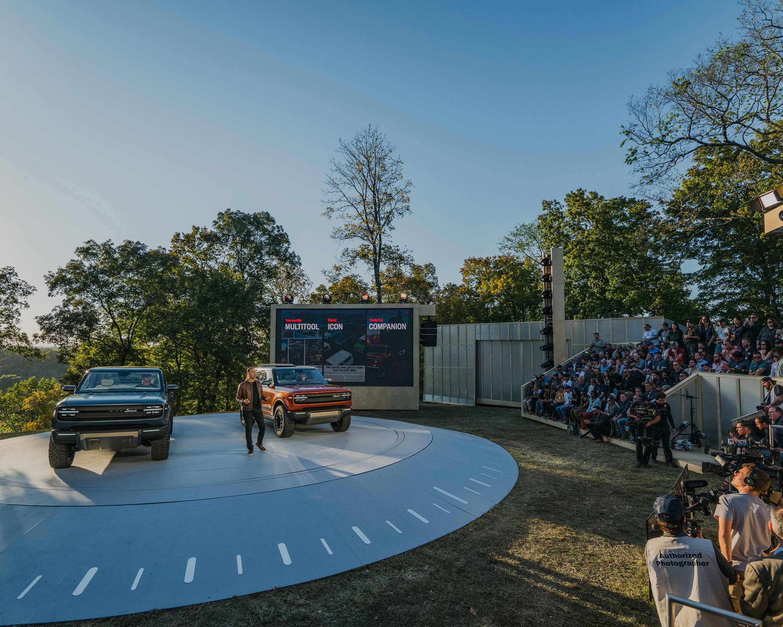 Two Rivian trucks are showcased outdoors on a circular platform for a large seated audience on a sunny day, surrounded by green trees.