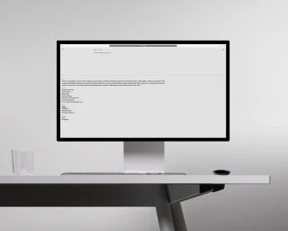 Minimalist computer monitor displaying website design on a sleek modern desk alongside a slim keyboard, mouse, and clear glass.