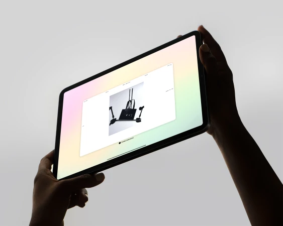 Close-up of an illuminated tablet with an interior design displayed on the screen, held by two silhouetted hands, neutral background.