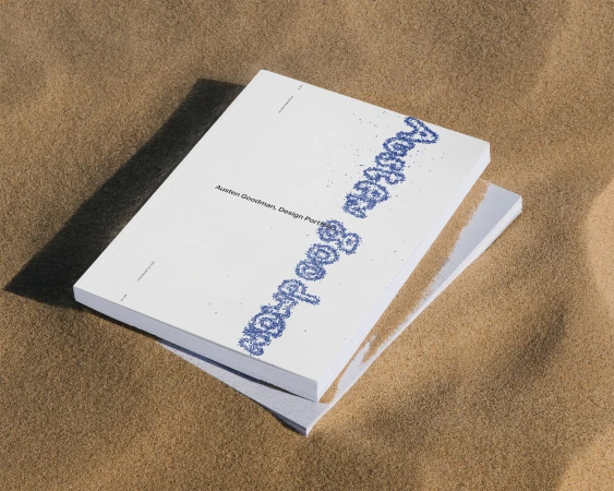 Stack of white books with blue stylized text on a sand surface in an outdoor setting with bright, diffused lighting.