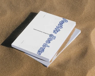 White design portfolio book with textured blue lettering resting on natural sand under bright sunlight.