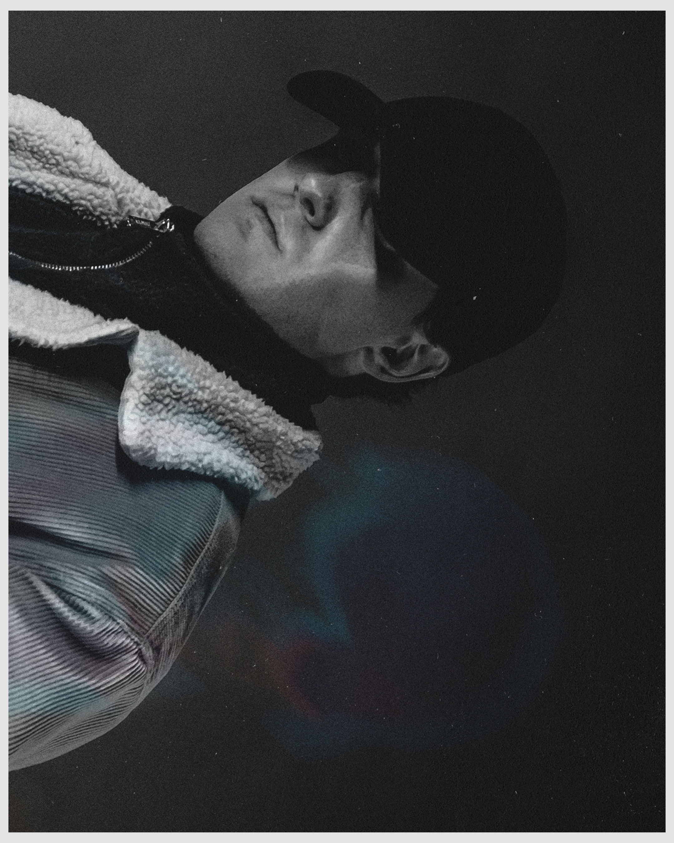 Portrait of a person wearing a black cap and denim jacket with a sherpa collar in a monochrome color palette and indoor setting.