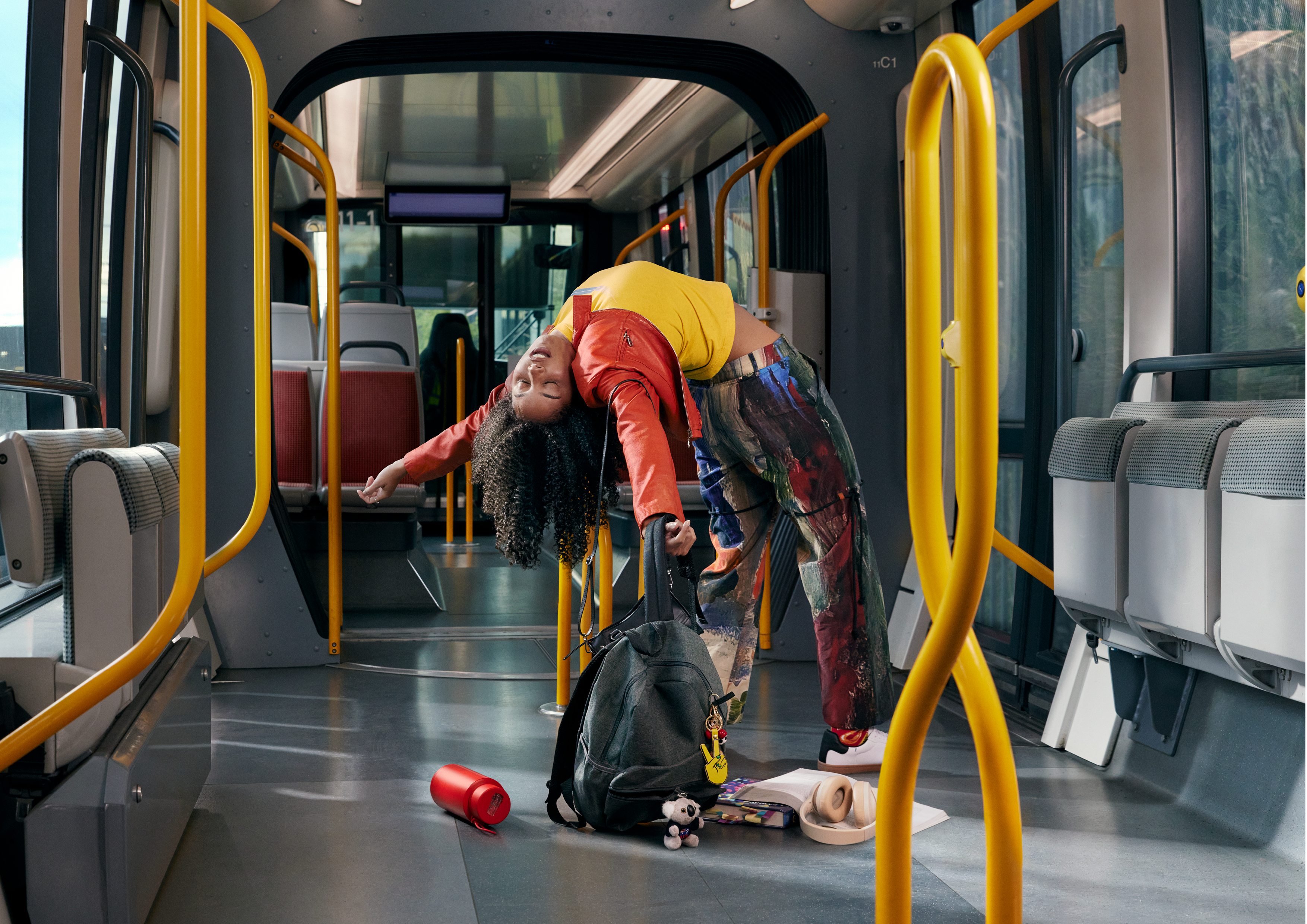A young woman bends over backwards on a bus with yellow handrails, wearing a yellow shirt, red jacket, and paint-splattered pants.