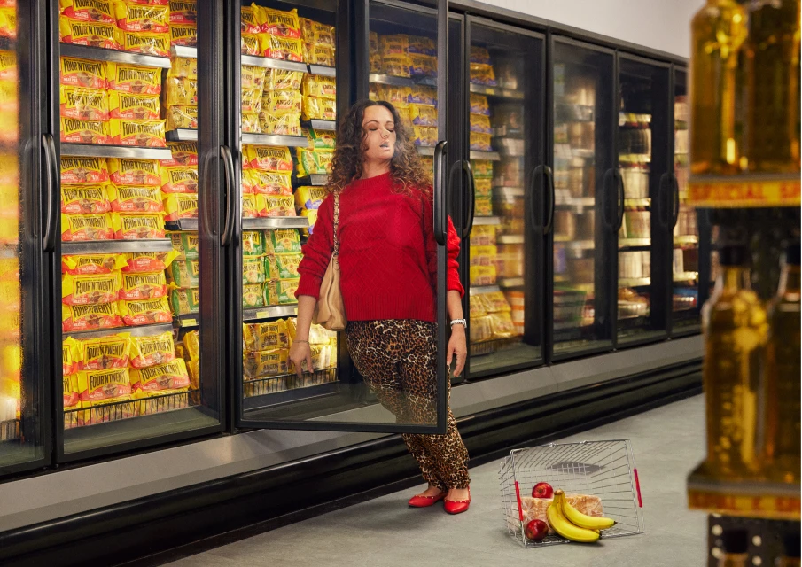A woman in a red sweater and leopard-print pants stands in front of a grocery store refrigerator holding a shopping basket with food inside.