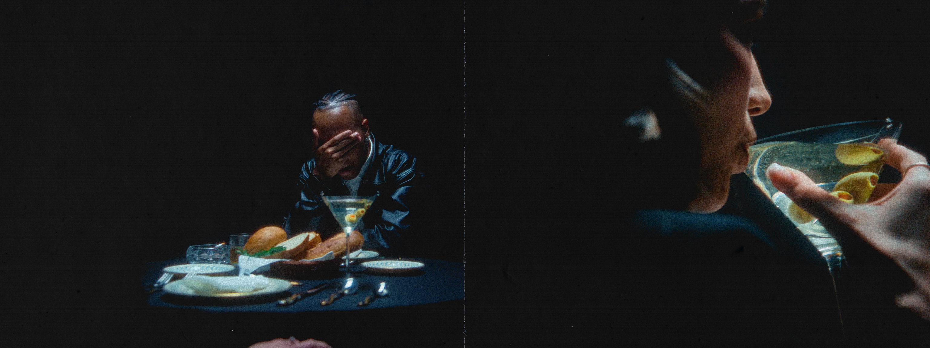 Man with braided hair sits at a dark table with a martini, bread, and dinnerware. A person sips a martini in a dark background.