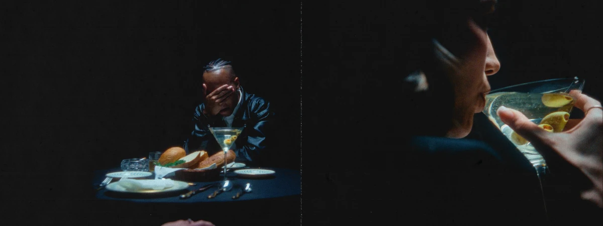 Man with braided hair sits at a dark table with a martini, bread, and dinnerware. A person sips a martini in a dark background.