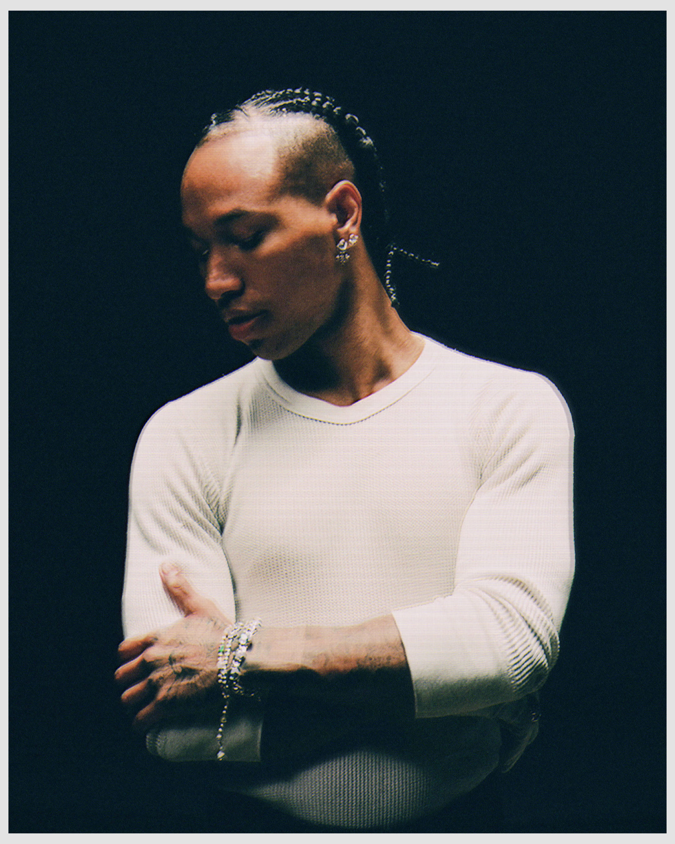 Portrait of a bald biracial male model with braided hair and jewelry, posing indoors against a black background in a white long-sleeved shirt.