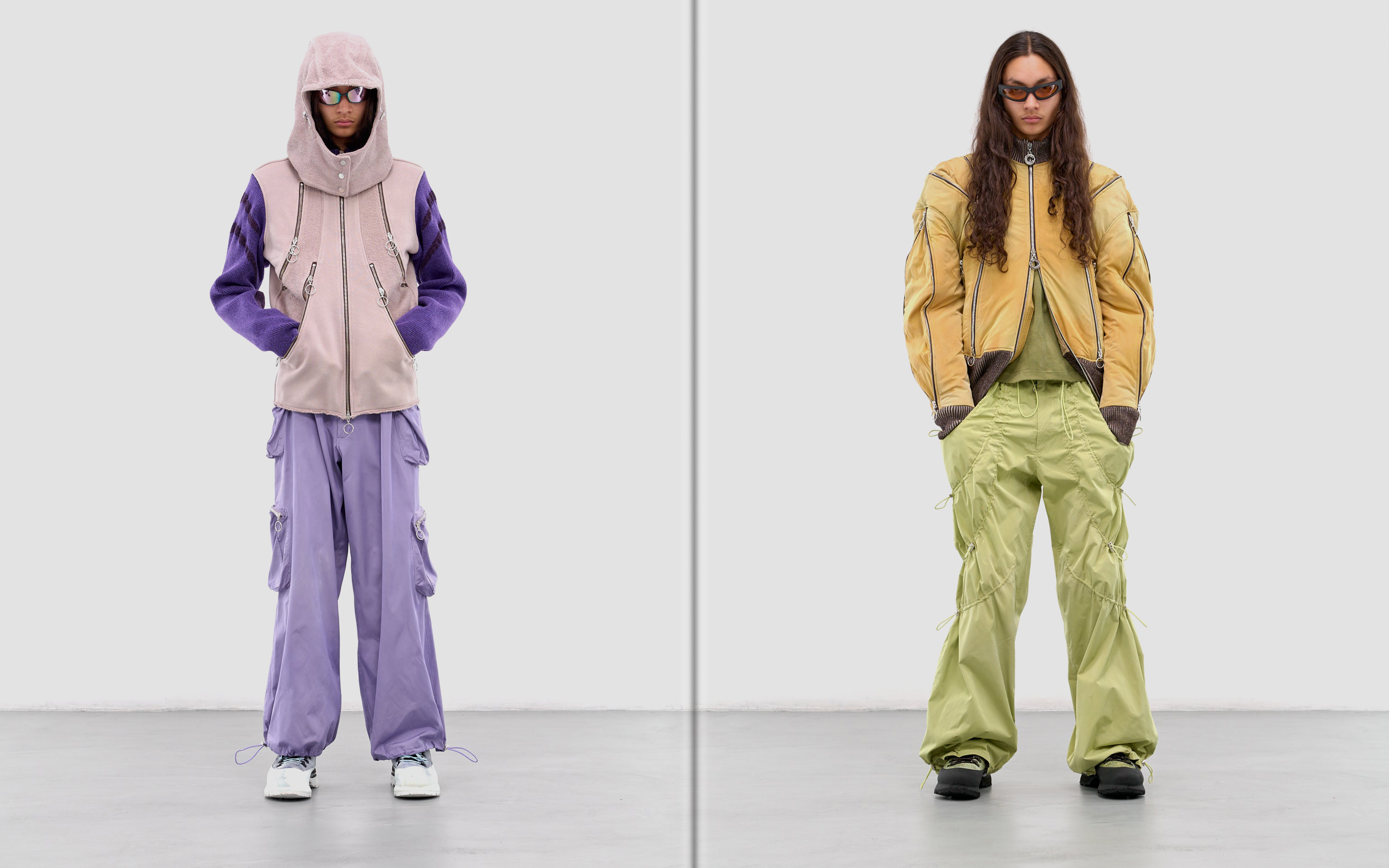 Two fashion models showcase avant-garde streetwear in a studio setting, featuring oversized jackets, cargo pants, and sunglasses against a plain backdrop.