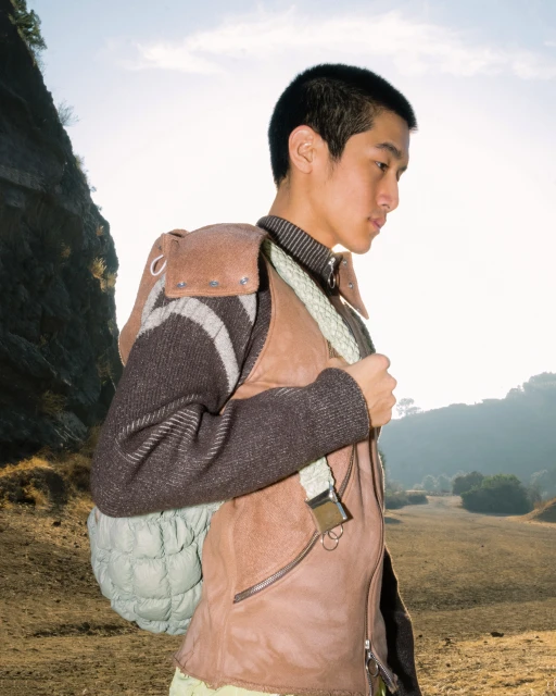 Young Asian man in outdoor field wearing vest and backpack, set against a backdrop of mountains and clear sky. Profile shot.