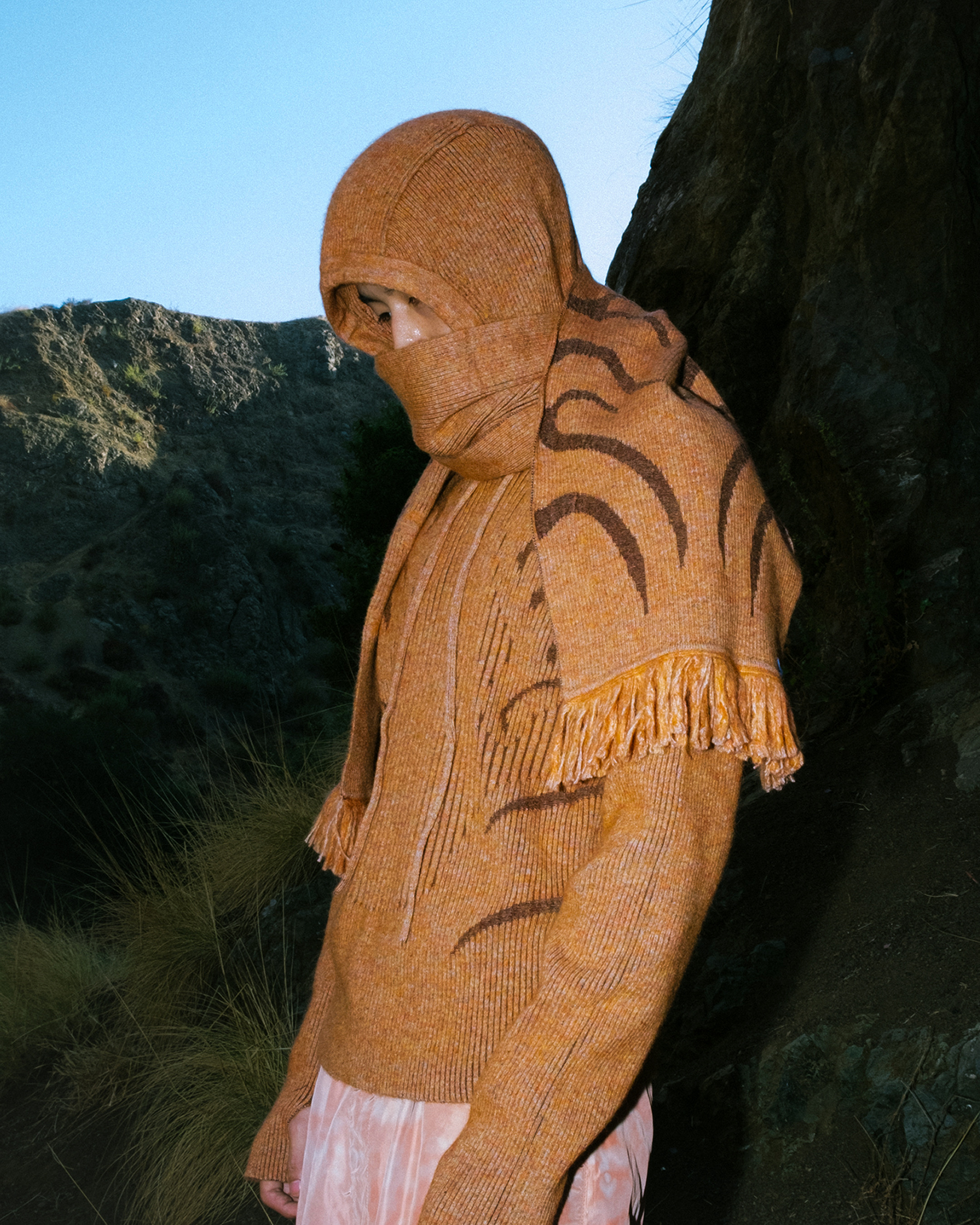 Person wearing orange knit hooded sweater and patterned scarf outdoors against a background of mountains and sky.