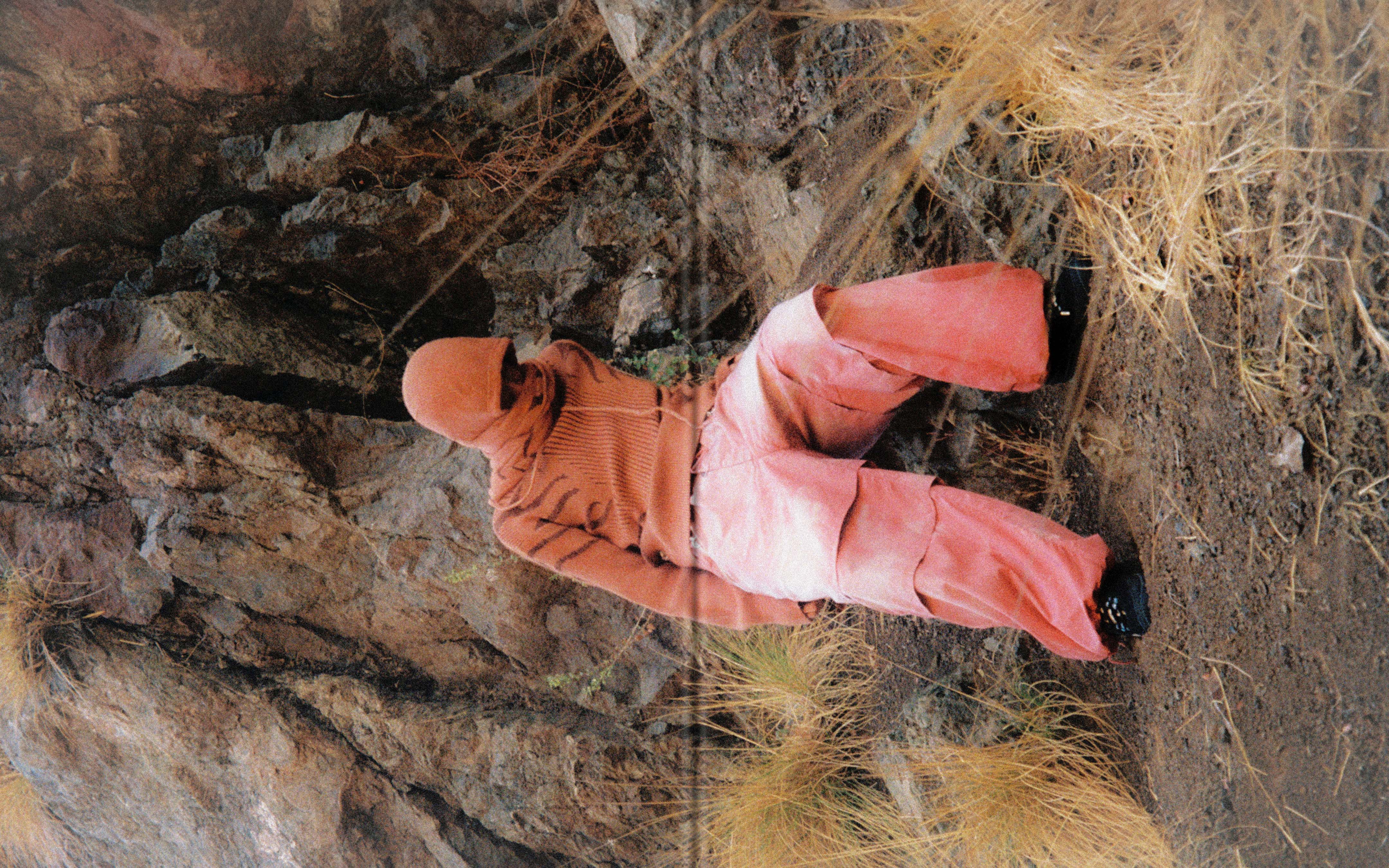 An adult poses wearing red, hooded fashion in a rocky, natural landscape. The photo has a high angle and the subject is lying down.