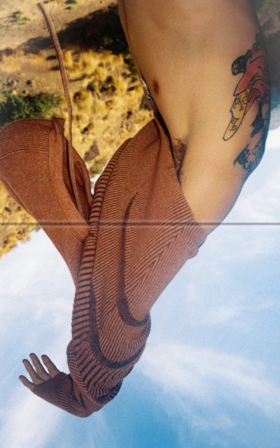 Conceptual studio shot of bare-chested person in knit sweater against an outdoor background of blue sky, clouds, and foliage.