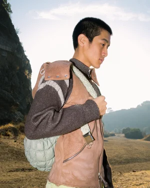 Man wearing a brown vest and knitted sweater with a light green quilted backpack outdoors against a rocky landscape