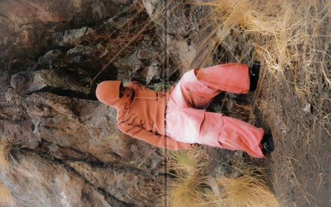 Model in orange hooded top and red trousers crouching against rocky cliff background