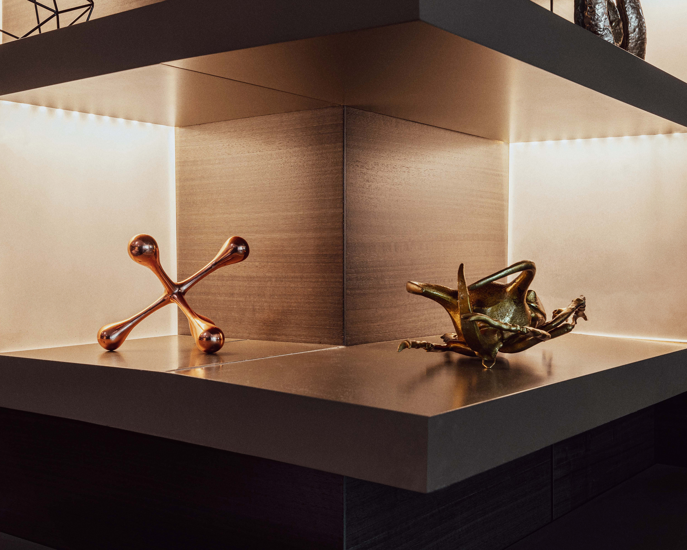 Corner shelf with bronze decorative objects. One is a jack-like shape and another is a sculptural piece; warm lighting highlights textures.