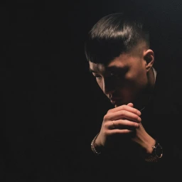 Portrait of a man with short dark hair and a watch holding his hands together in low-key lighting against a dark background