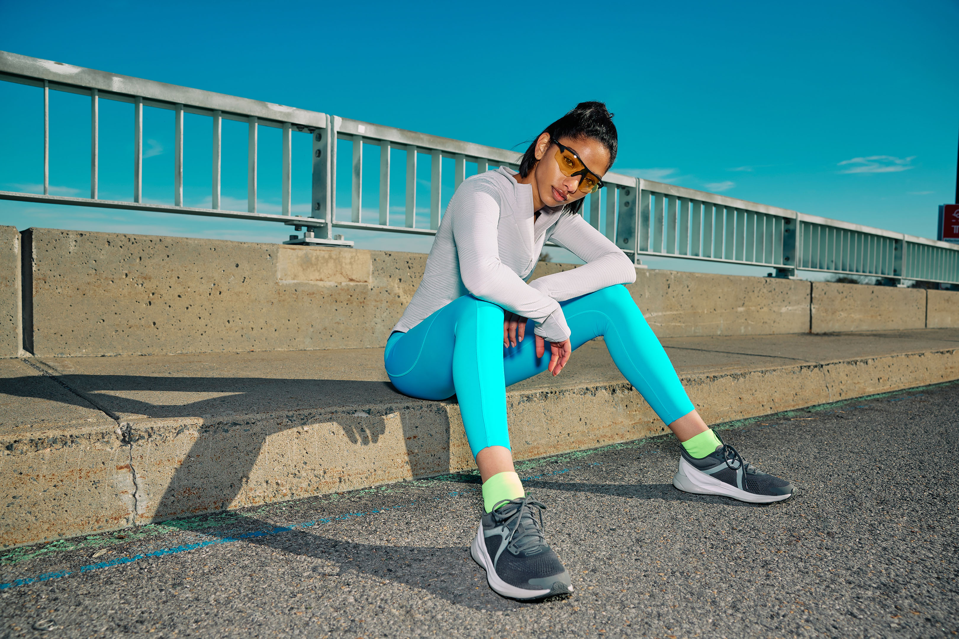 A young woman with sunglasses and athletic wear sits on concrete steps against a clear blue sky in an urban outdoor setting.