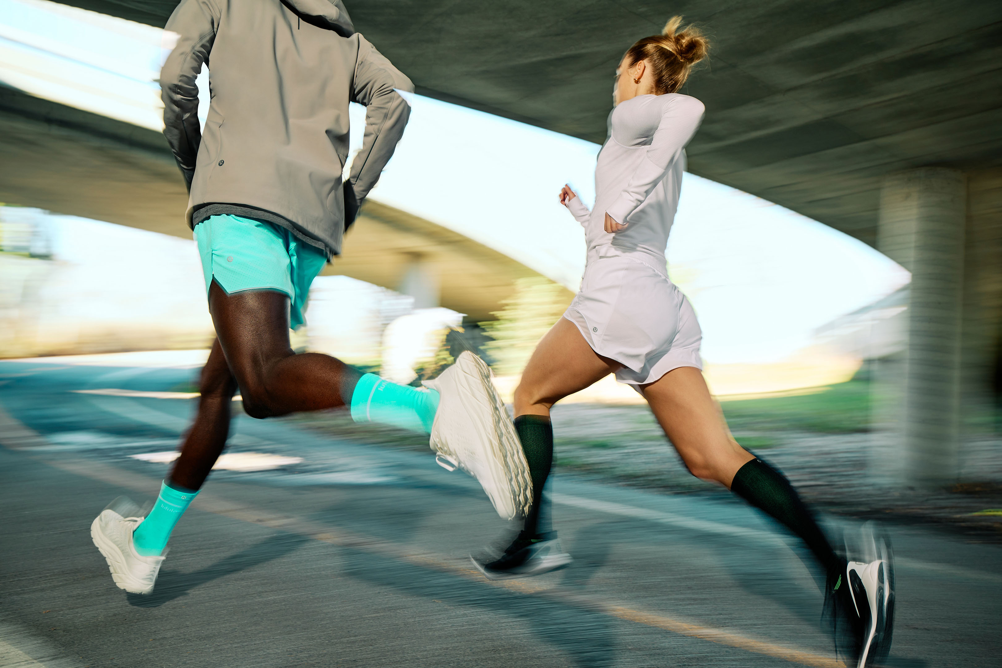 Two athletes, one Black and one white, are captured in motion running on asphalt near a concrete structure, with a blurred background.