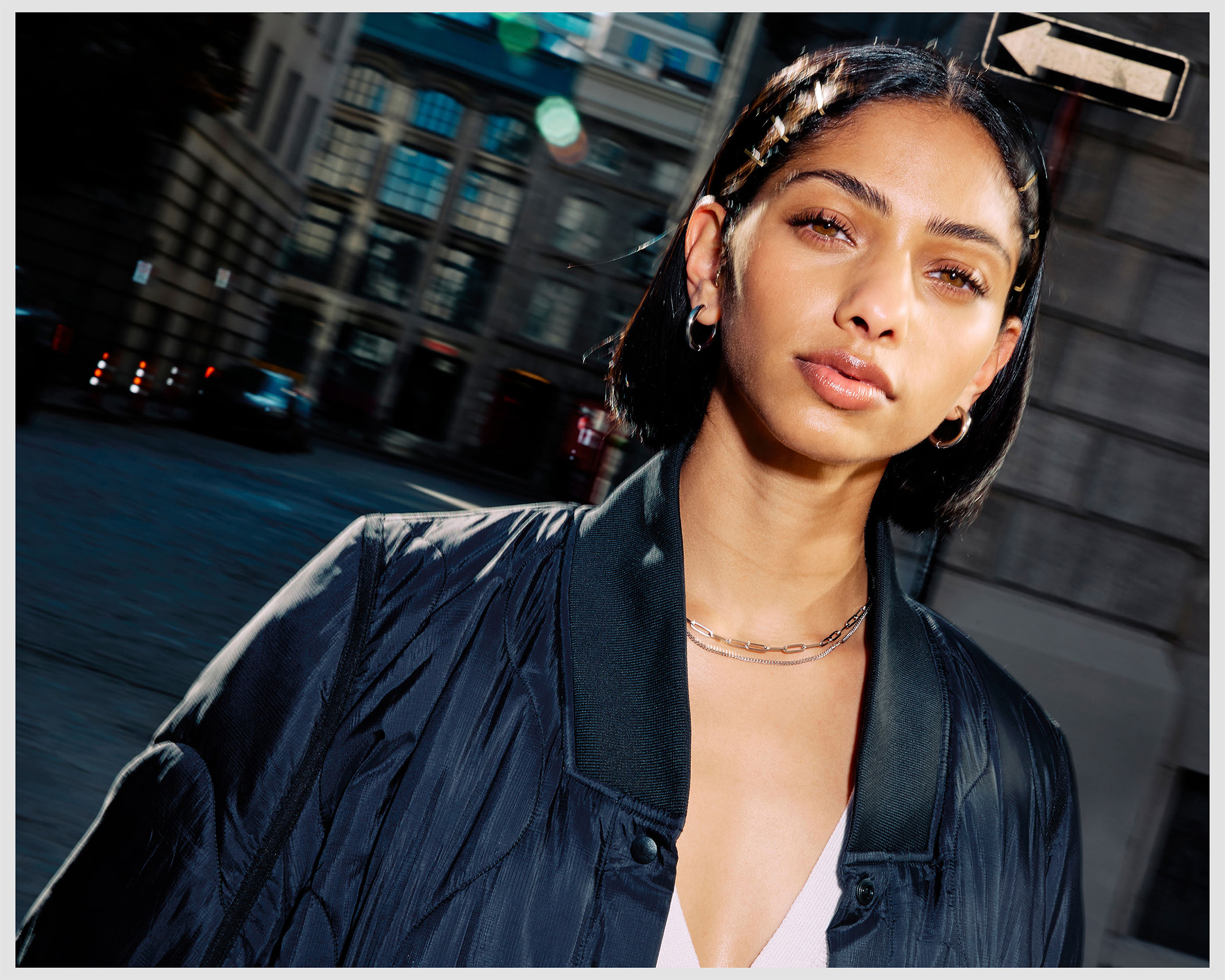 Portrait of a young adult woman with dark hair and braids, wearing a navy jacket and hoop earrings, stands in an urban street setting.