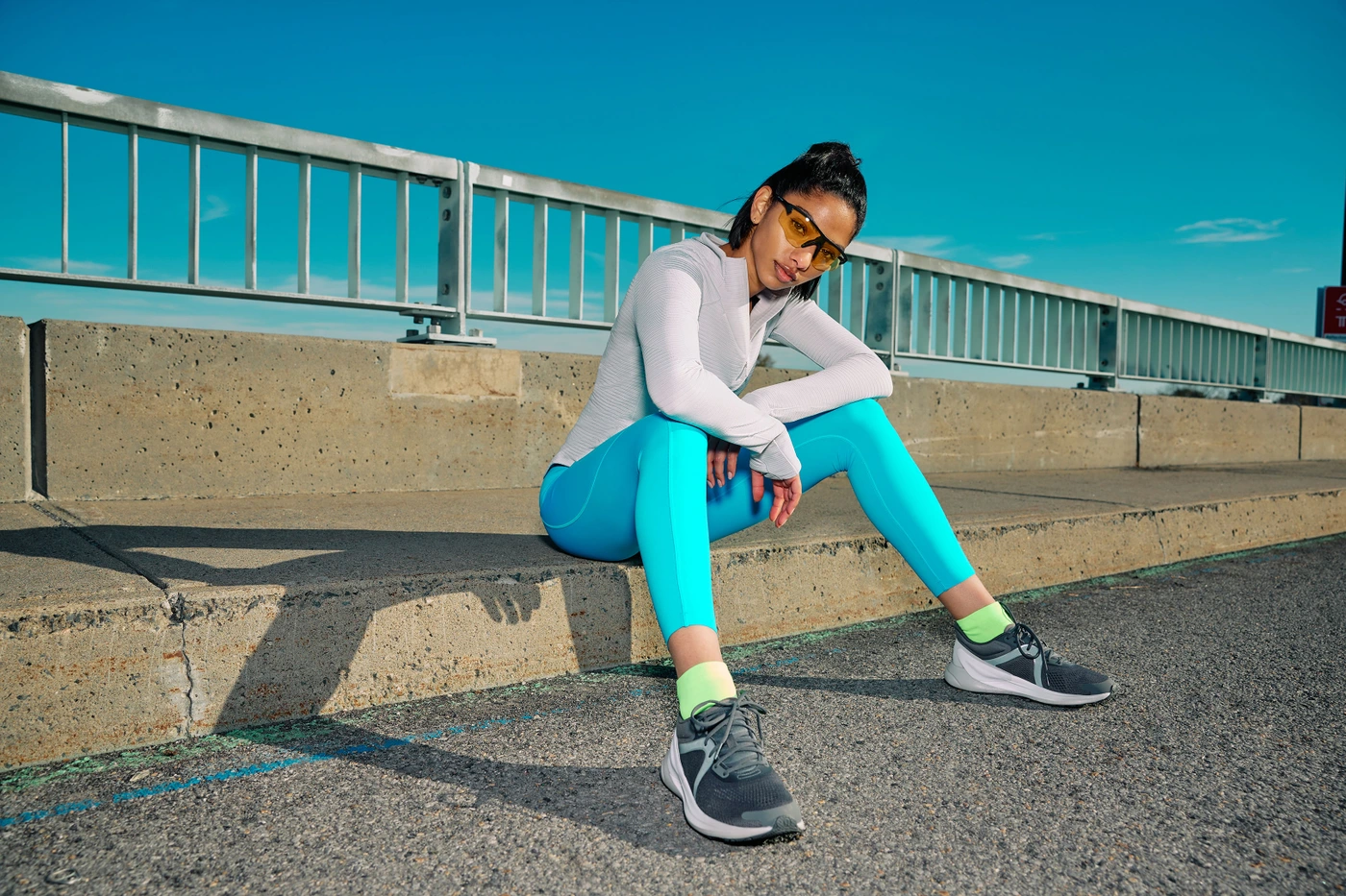 Woman in cyan leggings, grey long-sleeve top, and yellow sunglasses sitting on a concrete barrier along a bridge railing.